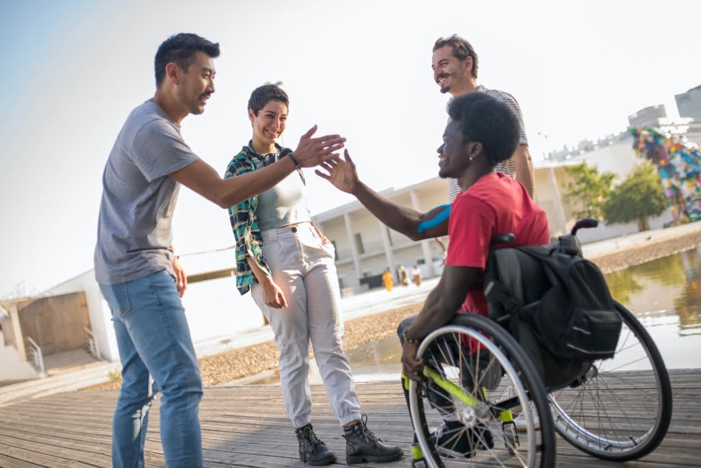 A diverse group of friends from different cultures in a group. A young Black man in a wheelchair is about to receive a handshake.