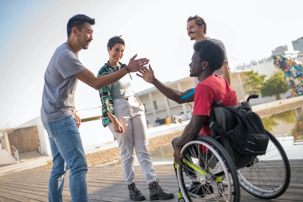 A diverse group of friends from different cultures in a group. A young Black man in a wheelchair is about to receive a handshake.
